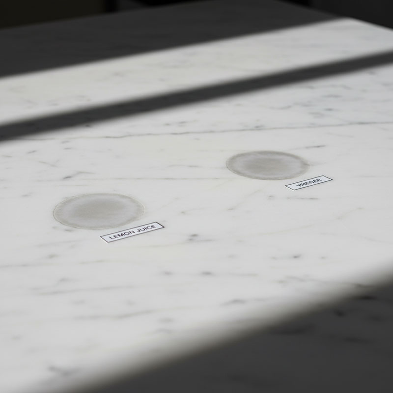 Close-up photograph showing acid etching damage on polished white marble surface with visible dull spots and rings from lemon juice and vinegar contact test
