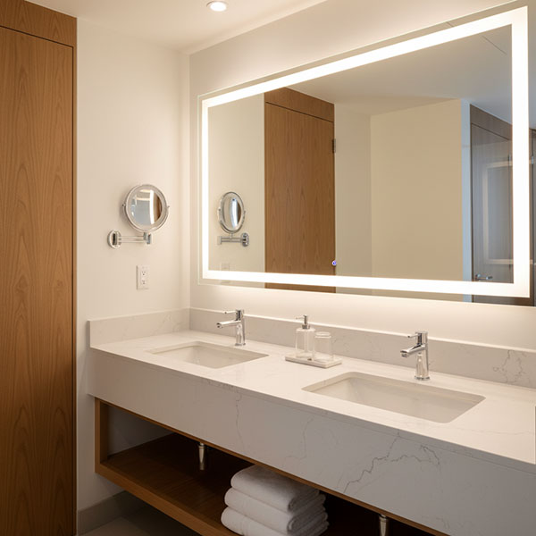 Modern luxury hotel bathroom interior with marble-look engineered quartz vanity countertop, frameless mirror, warm LED lighting, and clean contemporary design in white and grey tones